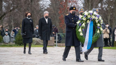 Presidentparet Sauli Niinistö och Jenni Haukio deltar i kransnedläggning vid Hjältekorset på Sandudds begravningsplats i Helsingfors 2020. Presidentparet har båda svarta ytterkläder och munskydd på sig. Framför dem bär tre män kransen.