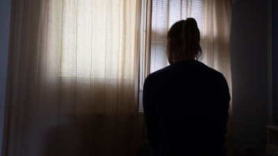 En ung kvinna sitter i ett mörkt rum med gardinerna fördragna.