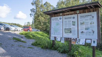 En skylt som det står Byabäcken på och innehåller infromation som Sibbo storskog. I bakgrunden ser man bilar på en parkeringsplats.