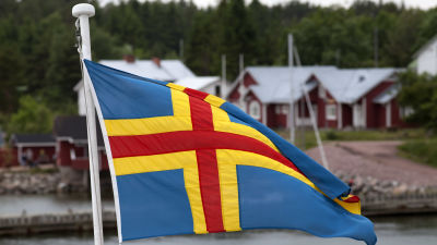 En åländsk flagga i förgrunden, röda hus med vita knutar i bakgrunden.