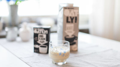 Ett glas med havredryck står framför en literburk med havremjölk. I bakgrunden syns också havregrädde.