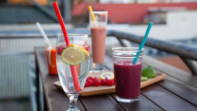 Glas med sugrör i står på ett bord. Glasen har olika saker i, t.ex. smoothie och vatten. På bordet finns också en bricka med frukt.