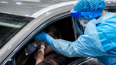 Coronatestning där person i bil testas av vårdare i skyddsutrustning.