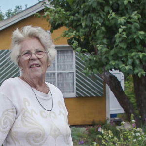 Vanhempi rouva vaaleassa puserossa ja mustavalkoruudullisissa housuissa seisoo siirtolapuutarhansa portilla. Taustalla keltainen mökki ja suuri omenapuu.