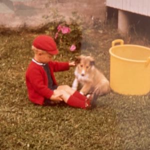 Punaista paitaa, nilkkasukkia ja hattua käyttävä poika istuu koiran kanssa nurmikolla. Taustalla iso vesiämpäri.