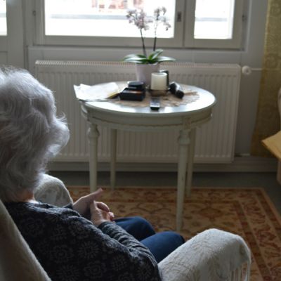 Vanha nainen istuu nojatuolissa ja katsoo ikkunasta ulos olohuoneessa.