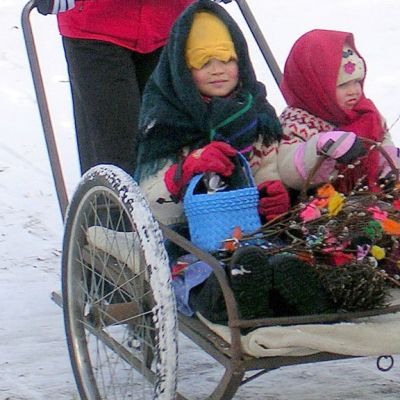 Kaksi tyttöä trulliasuissaan maitokärryissä. Vierellä hinkki kädessä kulkeva poikatrulli