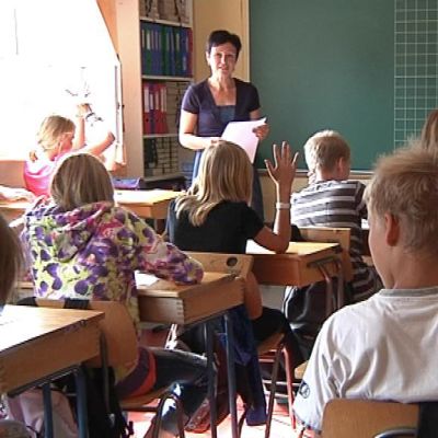 Opetustilanne koululuokassa, oppilaita viittaamassa, opettaja seisoo luokan edessä