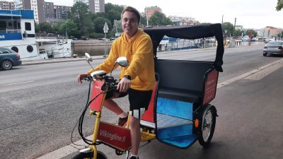 En ung man kör en gul cykeltaxi, riksha, längs en åstrand.