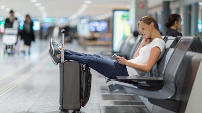 Kvinna sitter tillbakalutad på bänk i en terminal. Hon har fötterna på sin resväska och surfar på sin pekplatta.