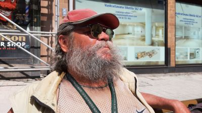 Markku Hynönen, en man med ett stort gråsvart skägg, solglasögon och en röd skärmmössa, sitter framför en byggnadsställning.