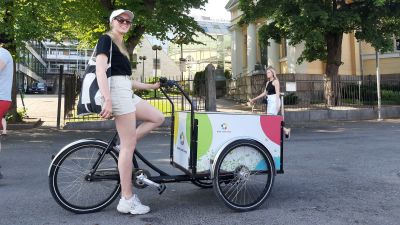 En dam med solglasögon, keps och shorts sitter på en flakcykel med färggranna bollar på lådan.