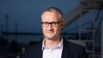Puvuntakkiin ja kauluspaitaan pukeutunut mies katsoo kameraan varovaisesti hymyillen taustanaan hämärä satama