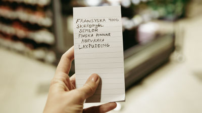 En hand håller i en uppköpslista i en mataffär. Listan räknar upp "fransyska 400 gram, skrädmjöl, semlor, finska pinnar, korvkaka, laxpudding".