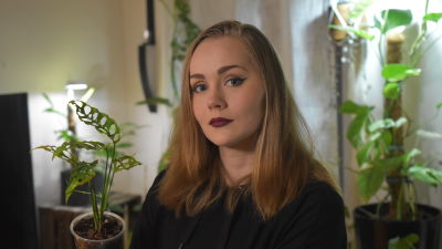 porträtt på en kvinna med axellångt hår, rött läppstift och vingad kajal. I bakgrunden syns krukväxter och i handen håller hon en monstera obliqua.