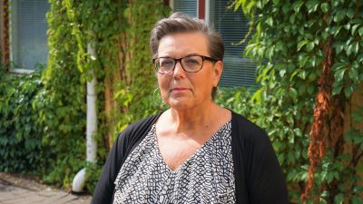 Kvinna med kort hår och glasögon står framför en gul byggnad.