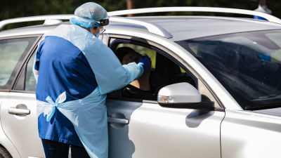 En person i skyddsutrustning tar ett coronatest på en person i en bil.
