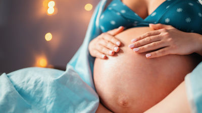 En gravid kvinnas sitter på en säng och håller i sin mage. I bakgrunden syns ljusbelysning.