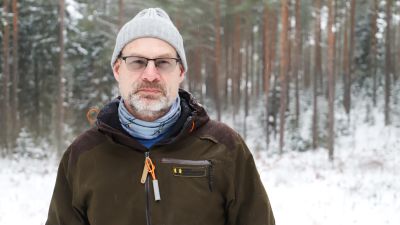 En man med grå mössa, glasögon och en mörkbrun rock står i en snötäckt skog på vintern.