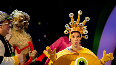 Kuvan etualalla on keltaiseen avaruusolentopukuun pukeutunut näyttelijä, jolla on päässään asuun kuuluva hattu, josta nousee lukuisia varrellisia pehmosilmiä. Koira-asuun pukeutunut näyttelijä vasemmalla virnistää. 