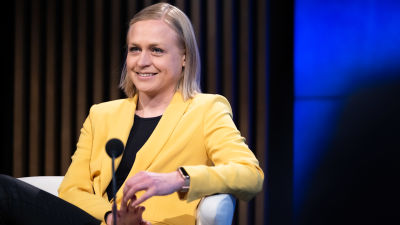Elina lepomäki, en blond kvinna klädd i gult, sitter leende i en vit stol.