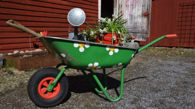 En grön skottkärra dekorerad med vita blommor, lastad med en hink med vita vårblommor, en vattenkanna i zink och en grep.