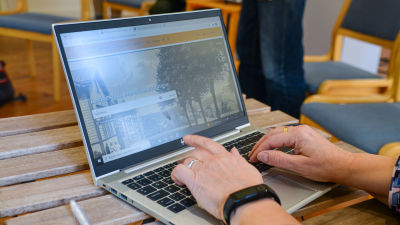 Händer skriver på tangentbordet på en laptop. På skärmen syns Lovisa stads museums nättjänst. Datorn står på ett litet träbord inomhus. 