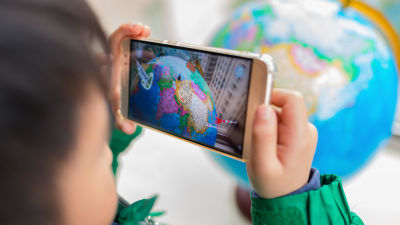 Kameran fokuserar på en mobilskärm som visar ett spel med en jordglob. I förgrunden ser vi ett suddigt barn som håller i mobilen.