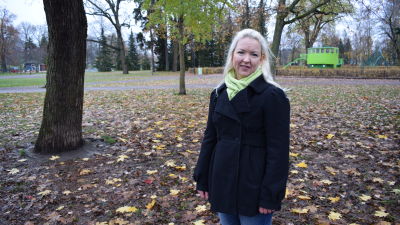 Kaisa Luntamo, en dam med blont långt hår och svart kappa, står i en park med kala träd och löv på marken.