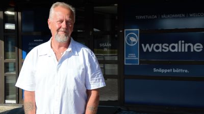 En skäggig man i vit skjorta står i solsken framför en färjeterminal.