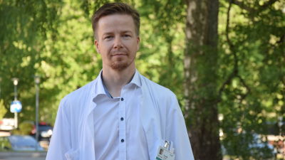 En man (läkare) i vit läkarrock poserar utomhus. Ser rakt in i kameran. Sommar.