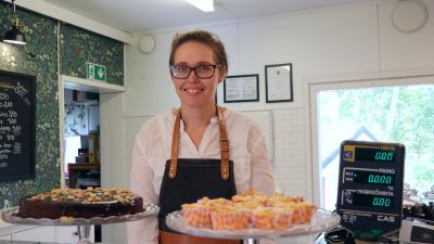 En kvinna iklädd vit skjorta och förkläde står bakom en disk i en butik med bakelser på fat i förgrunden.