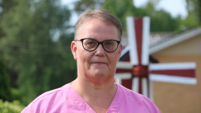 En medelålders kvinna med glasögon och vårduniform står utomhus.