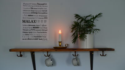 På väggen finns en plansch med ordet Malax och en mängd ord på Malaxdialekt. Under planschen finns en hylla. På hyllan står ett brinnande ljus och en grönväxt.