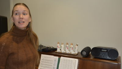 Inka-Maria Nyman, en dam med långt ljust hår och brun tröja, står framför att piano med luciafigurer och en radioapparat.