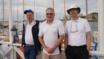Tre män står brevid varandra framför en hamn med segelbåtar.