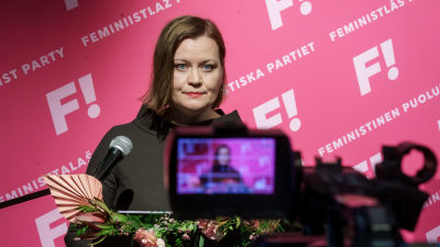 Katju Aro på valvaka med kamera i förgrunden och partireklam i bakgrunden.