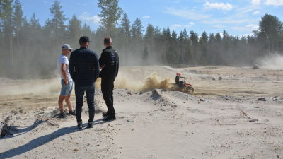 Tre män står och tittar på en motocrossförare som kör.