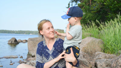 Sandra Söderholm sitter på en sten på en strandkant och håller om sin son Kevin Blomqvist.
