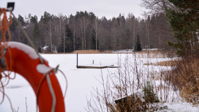 I förgrunden till vänster en röd livboj. Längre bort syns en träbrygga som leder ut i en frusen sjö i ett snöigt landskap. 