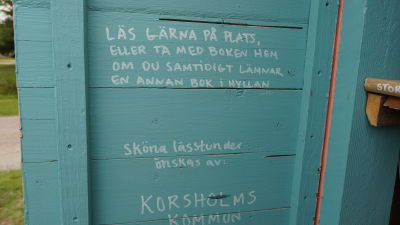 Ett turkost plank med vit text där det står: "Läs gärna på plats eller ta med boken hem om du samtidigt lämnar en annan bok i hyllan.