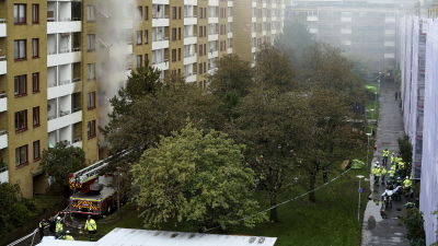 Bild på flervåningshus där det brinner. På marken står det flera brandmän.