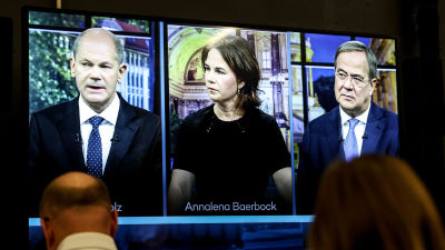 Tre tyska politiker sida vid sida på en tv-skärm. I förgrunden personer som tittar på skärmen.