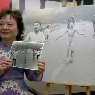 Kim Puc pitää kädessään kuvaa itsestään ja muista juoksevista lapsista, joiden takana kävelee Yhdysvaltain sotilaita. Taustalla on suurennos kuvasta.