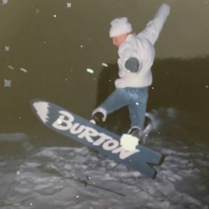 Farkkuihin ja valkoiseen talvitakkiin pukeutunut teini-ikäinen poika lumilautailee lumihangessa. Laudassa lukee Burton.