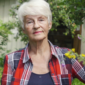 Vanhempi rouva ruutupaidassa seisoo toinen käsi puuskassa keskellä vehreää puutarhaa, päättäväinen ilme kasvoillaan.