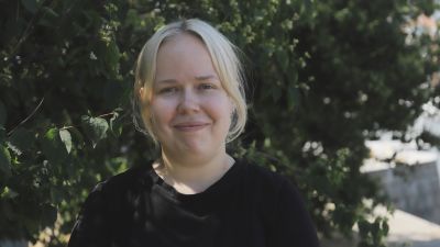 En ung kvinna med ljust hår står utomhus med en grönskande buske i bakgrunden.