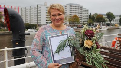 Radioredaktören Maisku Holmström blev vald till årets positivaste Åbobo. Bilden är fotograferad ombord på båten Ukko-Pekka I bakgrunden Aura å, i handen håller Holmström ett diplom och en blombukett.