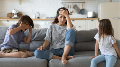 Trött mamma sitter på soffan med lekande barn.