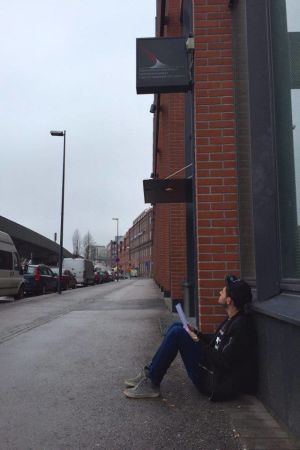 En ung man sitter på trottoaren med papper i famnen. Han tittar upp på Migrationsverkets skylt.